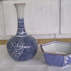 May include: A blue and white ceramic vase with a rounded body and a narrow neck. The vase has a geometric pattern with a floral design. A matching blue and white ceramic bowl with a hexagonal shape is also pictured.