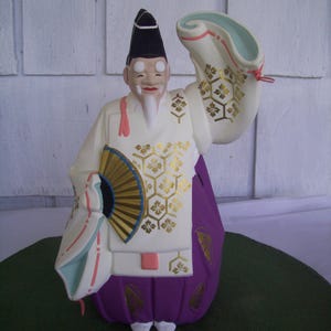 May include: A ceramic figurine of a Japanese man wearing a white and purple robe with gold accents. He is holding a fan and has a white beard.
