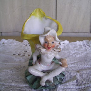 May include: A porcelain figurine of a whimsical character sitting on a green and yellow leaf. The character is wearing a white hat and a white outfit with red accents. The figurine is holding a small object in its hand.