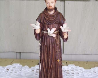 May include: A brown resin statue of Saint Francis of Assisi holding three white doves in his hands. The statue is standing on a brown base with the words "St. Francis" inscribed on it.