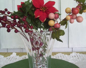 May include: A clear glass vase with a cut-glass design holds a bouquet of red poinsettia flowers, red berries, and pink and yellow berries.