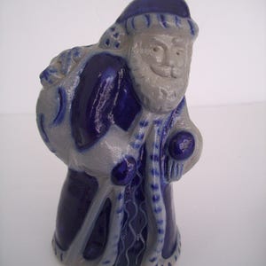 May include: A ceramic Santa Claus figurine in shades of blue and gray. The figure has a white beard and a blue hat. He carries a sack over his shoulder and is holding a small ball. The figurine is a decorative item.