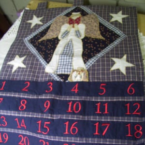 May include: A blue and white plaid fabric advent calendar with a quilted angel applique in the center. The calendar has numbers 1 through 24 embroidered in red on the fabric.
