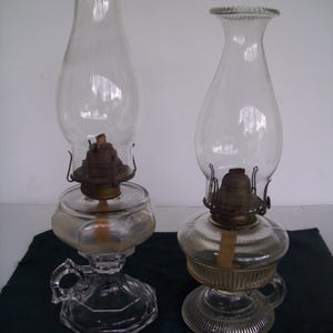 May include: Two clear glass oil lamps with chimneys. The lamps have a round base with a handle, a glass reservoir, and a metal burner. The chimneys are tall and cylindrical. The lamps are likely vintage or antique.