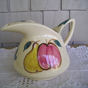 May include: A cream-colored ceramic pitcher with a pear and apple design. The pitcher has a spout and a handle. The pear is yellow with brown outlines and the apple is pink with brown outlines.