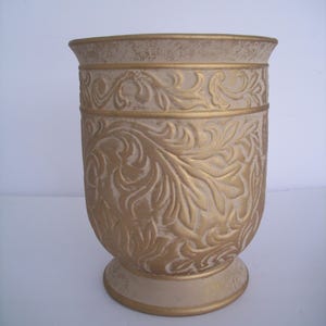 May include: A decorative vase with a cream-colored base and body, adorned with gold-colored, embossed floral patterns. The rim and bands are also gold, creating a classic, elegant look. The vase stands approximately 8 inches tall.