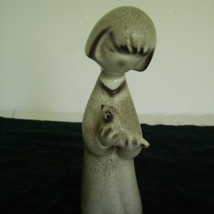 May include: A ceramic figurine of a person with a bob haircut, wearing a long robe with a speckled beige and brown finish. The figure is holding a small bird in both hands, and the base of the robe features a dark brown geometric border pattern.