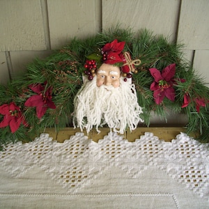 May include: A white, fluffy beard and red poinsettia flowers adorn a green pine garland. The garland is draped over a white crocheted tablecloth.