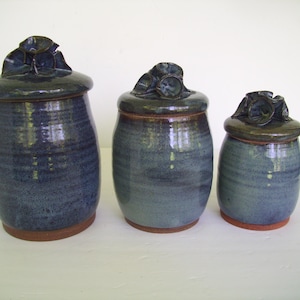 May include: A set of three cylindrical ceramic canisters with lids, each featuring a dark blue and grey mottled glaze. The lids are topped with sculpted floral-like handles, and the base of each canister reveals a natural, unglazed terracotta rim.