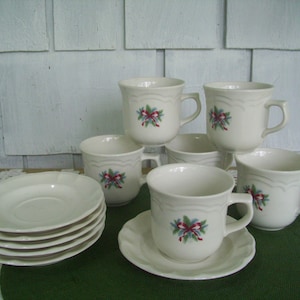 May include: Set of six white ceramic coffee cups with a green and red floral design. The cups are arranged in a circle on a green surface.