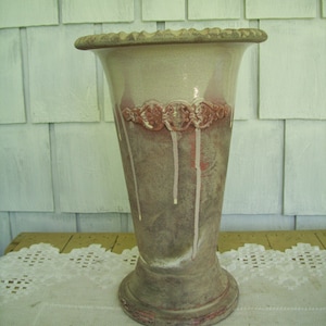 May include: A ceramic vase with a light brown glaze and a band of red and white around the middle. The vase has a flared top and a rounded base.
