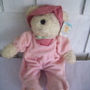 May include: A white plush teddy bear wearing a pink and red gingham sleepwear outfit. The bear has a red and white gingham hat and a tag that reads "Cuddle Me Toys".