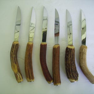 May include: A set of six steak knives arranged in a vertical row. Each knife features a polished, silver-toned metal blade and a curved, textured handle made from natural brown antler material, showcasing unique organic patterns and shapes.
