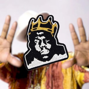 May include: A black and white embroidered patch of a person wearing a gold crown. The patch is in focus and the background is blurred.