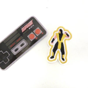 May include: A yellow and black embroidered patch of a video game character, Scorpion from Mortal Kombat, with a white background. The patch is next to a Nintendo Power mints container.