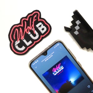 Puede incluir: Un parche bordado en blanco y negro con el texto "Wolf Club" en letras rosas. Un teléfono inteligente muestra una pantalla de reproductor de música con la portada del álbum "Wolf Club" de "All We Live For".