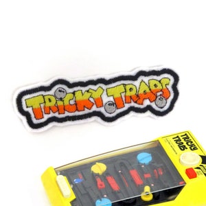Op de afbeelding: Een witte geborduurde patch met de woorden "Tricky Traps" in zwarte, oranje en gele letters. De patch heeft een zwarte omtrek en drie grijze ballen zijn geborduurd op de patch.