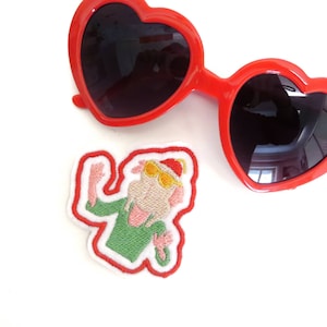 Op de afbeelding: Een geborduurde strijk-op patch met een cartoonfiguur dat een groen shirt, een rode hoed en een zonnebril draagt. Het figuur zwaait met één hand en heeft een roze gezicht.