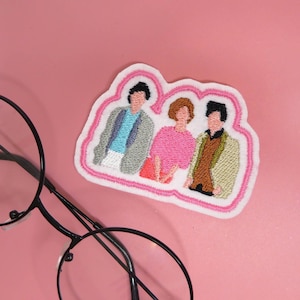 Op de afbeelding: Geborduurde strijkpatch met drie cartoonfiguren. De figuren zijn een man in een grijs jasje, een vrouw in een roze trui en een man in een bruin jasje. De patch is wit met een roze omtrek.