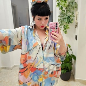 May include: A person wearing a colorful abstract print button-down shirt with short sleeves. The shirt has a light background with orange, blue, and yellow abstract shapes.