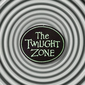 Può includere: Una toppa ricamata in bianco e nero con il testo "The Twilight Zone" in lettere verdi.