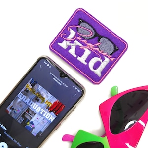 Puede incluir: Un parche bordado morado con la palabra "Kid" y unas gafas de sol. Un smartphone muestra la portada del álbum "Graduation" de Sunglasses Kid. También hay gafas de sol rosas y verdes brillantes en la imagen.
