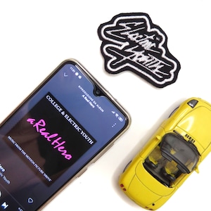 May include: A black smartphone displaying the song "a Real Hero" by College & Electric Youth from the movie "Drive". The song is playing on a music streaming app. The phone is on a white surface next to a yellow toy car and a black and white embroidered patch that says "Electric Youth".
