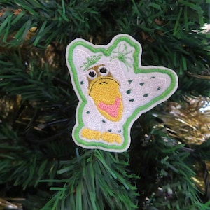 May include: A white embroidered patch with a green outline, depicting a cartoon bird with a pink beak and yellow feet. The bird is wearing a white hooded garment with green spots and has a green leafy crown on its head.