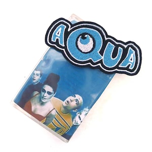 May include: A blue and black embroidered patch with the word "AQUA" and a blue eye with a white pupil. The patch is on top of a cassette tape case with a band photo on the front.