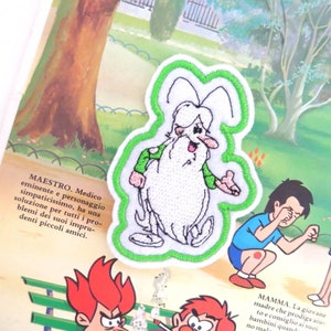 May include: Green embroidered patch of a cartoon character with a white beard and green shirt. The character is standing with a white background and green outline. The text "MAESTRO. Medico eminente e personaggio simpaticissimo, ha una soluzione per tutti i problemi dei suoi imprudenti piccoli amici." is visible in the background.