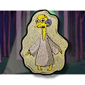 May include: An embroidered patch depicting a yellow cartoon character with a long neck and large eyes, wearing a grey robe. The character is surrounded by a white glow and is set against a dark background.