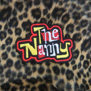 Può includere: Una toppa ricamata con il testo "The Nanny" in rosso, giallo e bianco su uno sfondo leopardato.