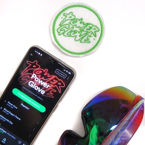Puede incluir: Un teléfono inteligente que muestra una aplicación de música con el nombre "Power Glove" en texto rojo neón. La aplicación muestra una lista de canciones populares, incluyendo "Power Core" y "Blood Dragon Theme". El teléfono está sobre una superficie blanca junto a un parche bordado verde y blanco con las palabras "Power Glove" en verde.