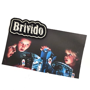 Könnte beinhalten: Ein gestickter schwarzer und weißer Aufnäher mit dem Wort "Brivido" in weißen Buchstaben. Der Aufnäher befindet sich auf einem Foto von drei Kindern, die ein Spiel mit einem Spukhaus spielen.