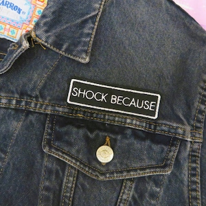 Könnte beinhalten: Dunkelblaue Jeansjacke mit gelben Nähten und einer Brusttasche. Ein schwarzer Aufnäher mit weißem Text "SHOCK BECAUSE" ist auf die Jacke genäht. Die Jacke hat einen silbernen Knopf an der Tasche.