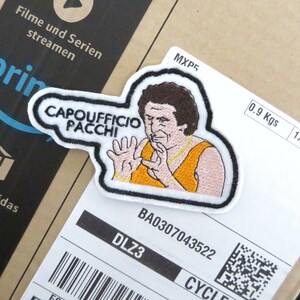 May include: White embroidered patch featuring a cartoon image of a man with brown hair and a moustache wearing a yellow tank top and a gold chain. The text "CAPOUFFICIO PACCHI" is written in black on the patch.