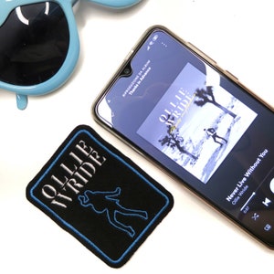 May include: A black patch with the text "OLLIE WRIDE" in white and a blue outline of a body. A smartphone displays the album art for "Never Live Without You" by Ollie Wride. Light blue sunglasses are in the background.