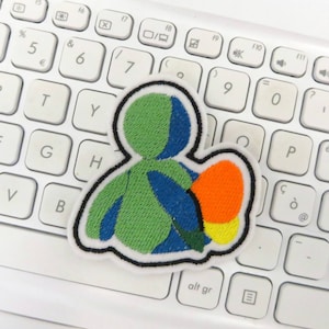Pode incluir: Patch bordado com uma figura estilizada com secções verdes e azuis, segurando uma forma laranja e amarela. O patch tem uma borda preta e está sobre um teclado branco.