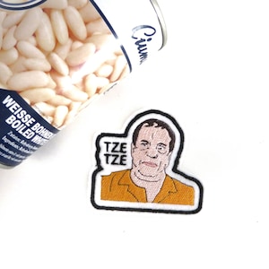 May include: An iron-on patch with an embroidered design. The patch features a cartoon portrait of a man with the text "TZE TZE" above him. The man is wearing a yellow shirt and has a serious expression.