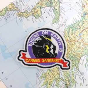 May include: A purple and white embroidered patch with a cartoon character wearing a hat and the text "Where on Earth is Carmen Sandiego?"
