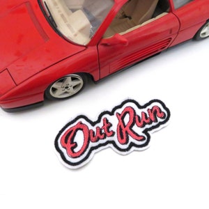 May include: A white embroidered patch with black outlining and red lettering that reads "Out Run".