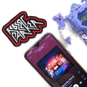 Puede incluir: Primer plano de un teléfono inteligente que muestra una aplicación de reproductor de música con la portada del álbum "Crystal City" de Robert Parker. La portada del álbum presenta un automóvil deportivo rojo conduciendo por una ciudad de noche. La canción "Again (feat. Miss K)" está sonando.