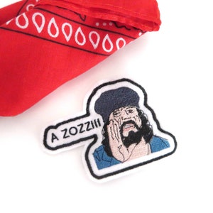 May include: A white embroidered patch featuring a cartoon man with a beard and a blue hat. The man is looking down and has his hand on his chin. The text "A ZOZZZI" is in a speech bubble coming from the man's mouth.