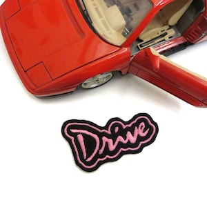 May include: A black and pink embroidered patch with the word "Drive" in a cursive font. The patch is on a white background.