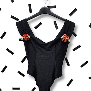 May include: Black ribbed bodysuit with a scoop neck and off-the-shoulder straps. The bodysuit has two orange and red fabric flowers on the shoulders.