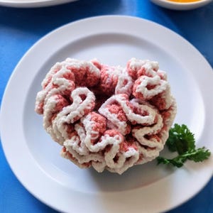 May include: A handmade crochet scrunchie in coral and white, resting on a white plate. The scrunchie has a textured, ruffled design. A sprig of parsley is next to the scrunchie. Other plates with food are in the background.