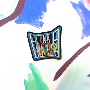 Könnte beinhalten: Ein blauer und schwarzer gestickter Aufnäher mit dem Text "CASA VANELLO" in bunten Buchstaben. Der Aufnäher ist in Form eines Fensters mit einem gelben Kreis in der Mitte.