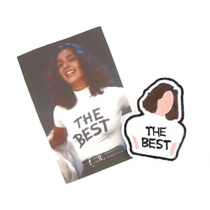 Peut inclure: Un t-shirt blanc avec le texte "THE BEST" en noir, et un écusson assorti. L'écusson présente une illustration stylisée d'une personne portant un t-shirt blanc avec le texte "THE BEST".