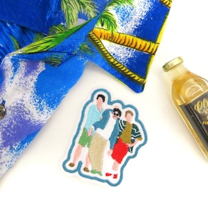 Può includere: Una toppa ricamata con tre figure di cartoni animati, due con camicie blu e una con una camicia verde. Le figure sono in piedi insieme e sorridono. La toppa è bianca con un contorno blu.