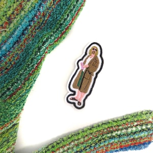 May include: Embroidered iron-on patch featuring a cartoon character wearing a brown robe and holding a green object. The character has long blonde hair and a beard.